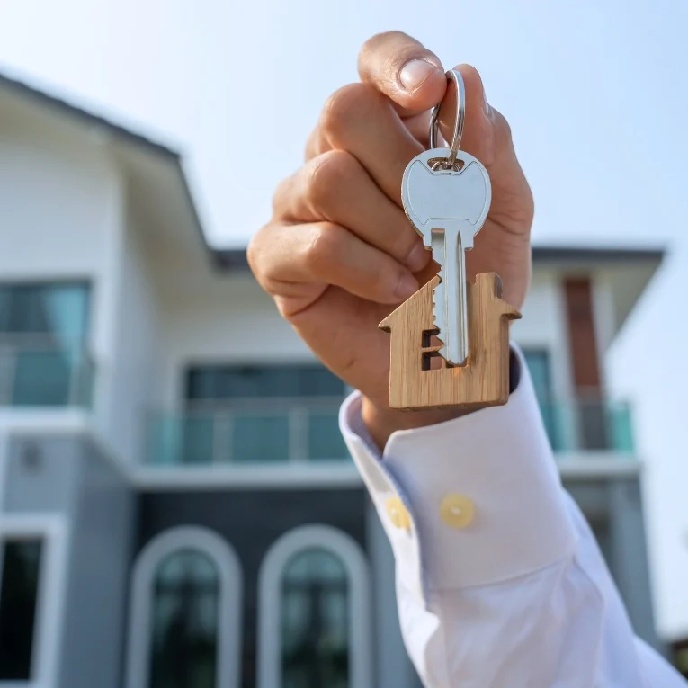 Hand holds house-shaped keychain in front of modern home, symbolizing new property ownership. Interior Design Wilmington, NC potential.